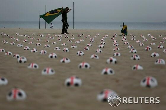 본문 이미지 - 브라질 리우데자네이루의 코파카바나 해변에서 22일(현지시간) 브라질 국기를 든 한 시위자가 빨간 십자가가 그려진 축구공들 사이를 걸어가고 있다. 이 축구공은 '리우 평화'라는 NGO에서 마련한 것인데, 교육, 의료, 공공서비스를 월드컵 경기장과 맞먹는 수준으로 해 줄 것을 정부에 요구하는 의미를 담고 있다.© News1 