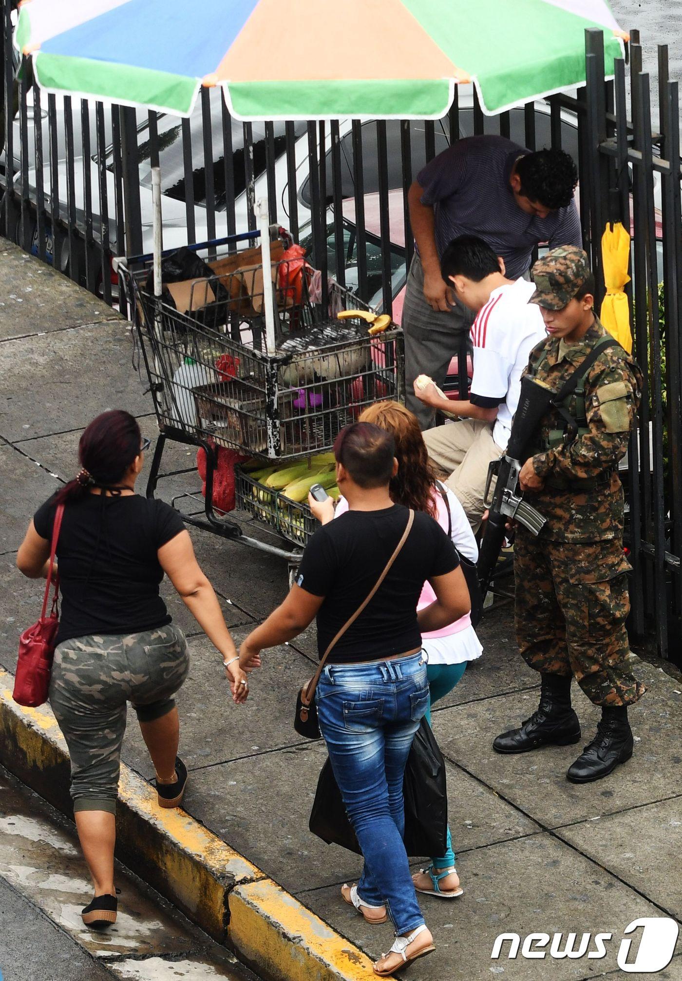 본문 이미지 - 엘살바도르 수도 산살바도르 시내 버스정류장에 배치돼 있는 순찰 군인 앞을 여성들이 지나가고 있는 모습. ⓒ AFP=뉴스1