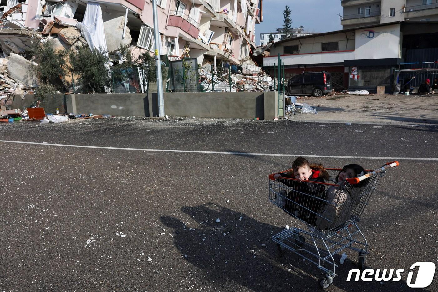 7일(현지시간) 튀르키예 하떠이 한 주택가에 두 꼬마가 시장 카트 속에 쪼그려 앉아 있다. 아이들의 뒤로는 강진으로 무너진 아파트와 잔해가 뒤죽박죽 섞여 있다. ⓒ 로이터=뉴스1 ⓒ News1 권진영 기자