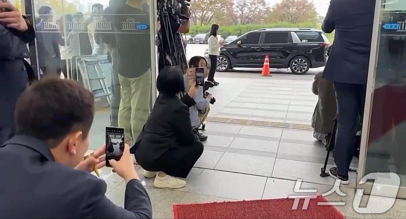 본문 이미지 - 그룹 뉴진스 멤버 하니(20·본명 하니 팜)가 15일 국회 환경노동위원회 국정감사장으로 참석한 가운데 국회 과학기술방송통신위원장인 최민희 더불어민주당 의원이 본청 입구에 쪼그리고 앉아 차에서 내린 하니를 바로 앞에서 촬영하고 있다. (이기인 개혁신당 최고위원 페이스북 캡쳐) 2024.10.15/뉴스1