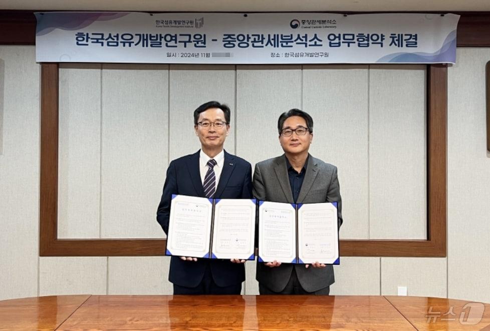 본문 이미지 - 호요승 한국섬유개발연구원장(왼쪽)과 박재선 중앙관세분석소장이 분석지원 및 보유 장비 공동 활용을 위한 업무 협약을 체결한 후 협약서를 펼쳐 보이고 있다.(섬유개발연구원 제공)