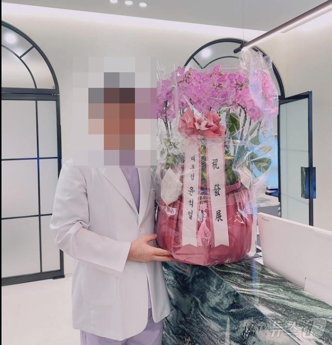 본문 이미지 -  대통령 자문의인 성형외과 전문의 박 모 원장이 지난해 8월 개업했을 때 윤석열 대통령 명의의 축하 화한을 옆에 두고 기념촬영을 했다. (SNS 갈무리) ⓒ 뉴스1