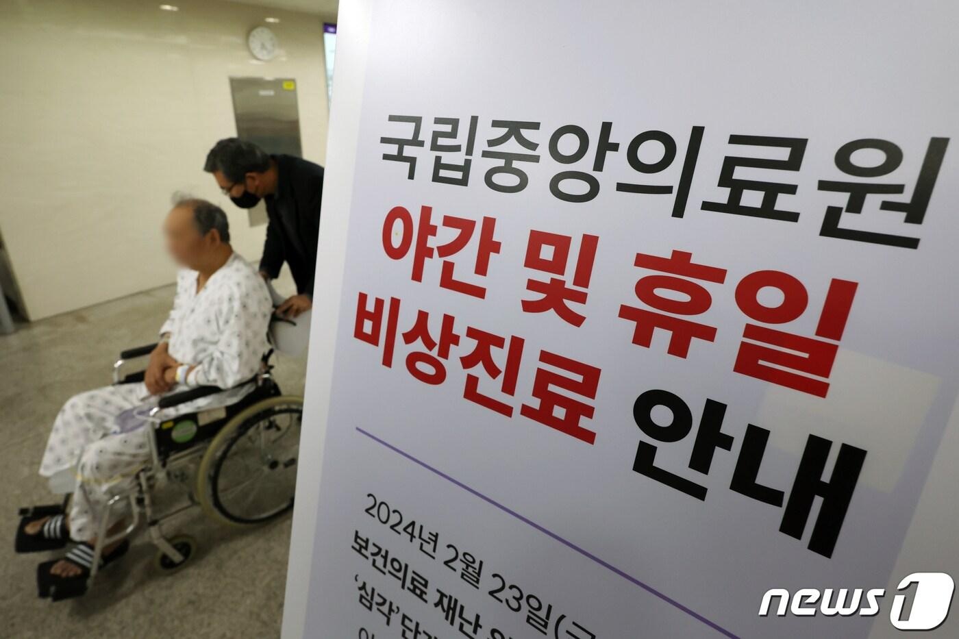본문 이미지 - 정부의 의대 정원 증원 방침에 반발한 전공의들이 집단행동이 일주일째 이어지면서 야간진료 공백이 우려되고 있다. 26일 저녁 서울 중구 국립중앙의료원에 게시된 야간 및 휴일 비상진료 안내문 뒤편으로 보호자가 환자의 휠체어를 밀며 걸음을 옮기고 있다. 2024.2.26/뉴스1 ⓒ News1 민경석 기자