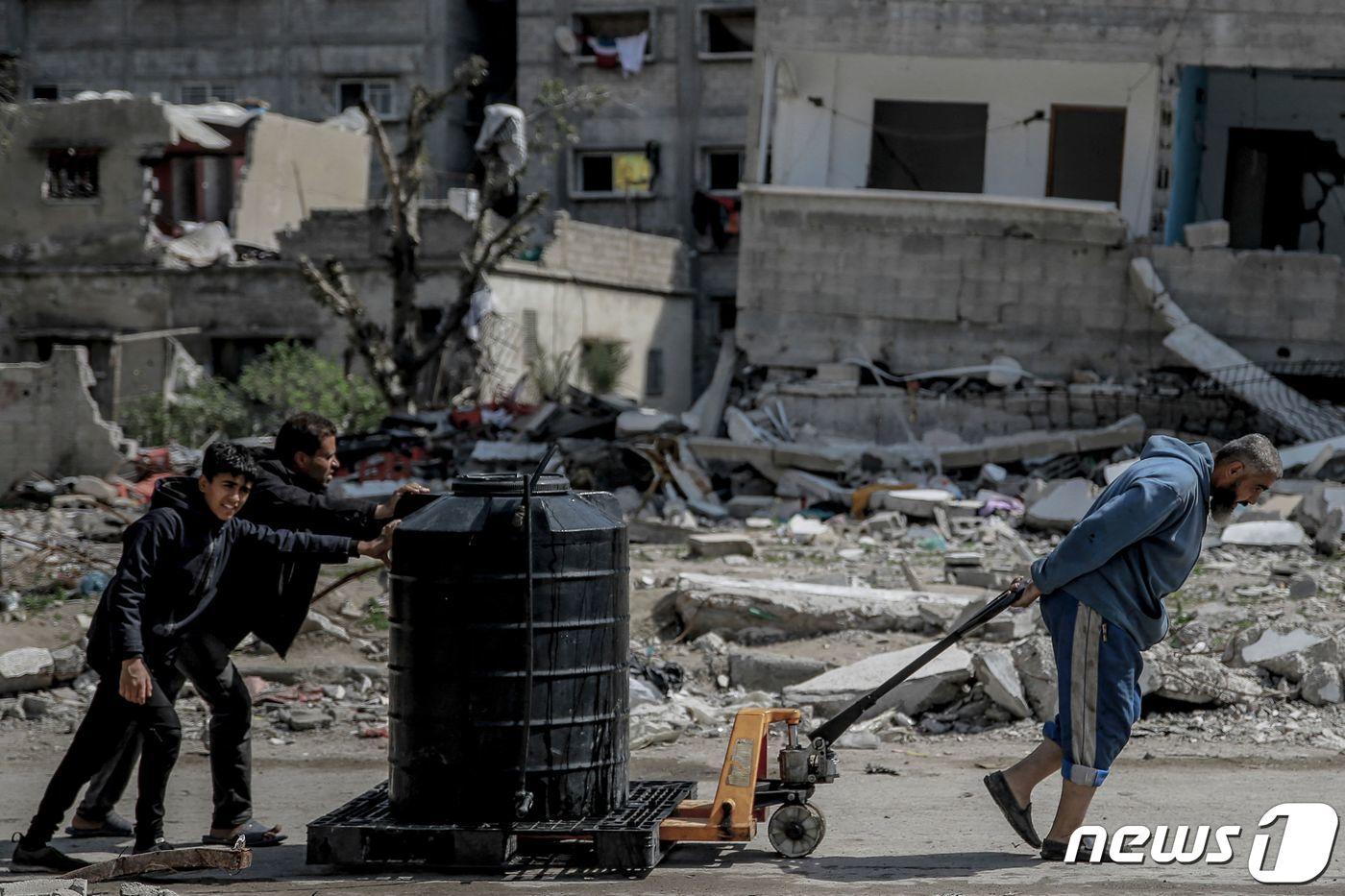 본문 이미지 - 3일(현지시간) 가자지구 한 거리에서 팔레스타인 남성과 소년들이 물탱크를 운반하고 있다. 배경에는 무너진 건물 잔해들이 보인다. 2024.03.03/ ⓒ AFP=뉴스1 ⓒ News1 권진영 기자