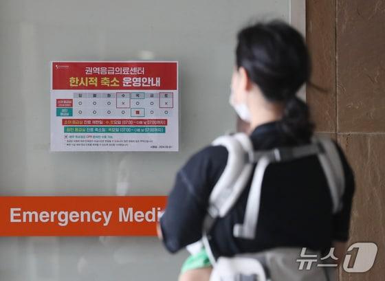 본문 이미지 - 소아를 안고 있는 한 보호자가 권역응급의료센터 한시적 축소 운영 안내문을 보고 있다./뉴스1 김영운 기자