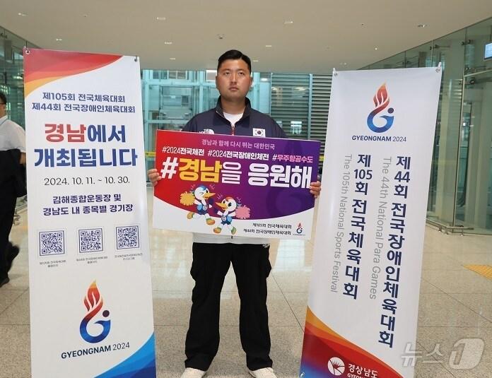 본문 이미지 - 양궁 김우진 선수가 경남에서 열리는 105회 전국체육대회를 응원하는 메시지가 담긴 피켓을 들고 전국체전을 홍보하고 있다.&#40;경남도 제공&#41;