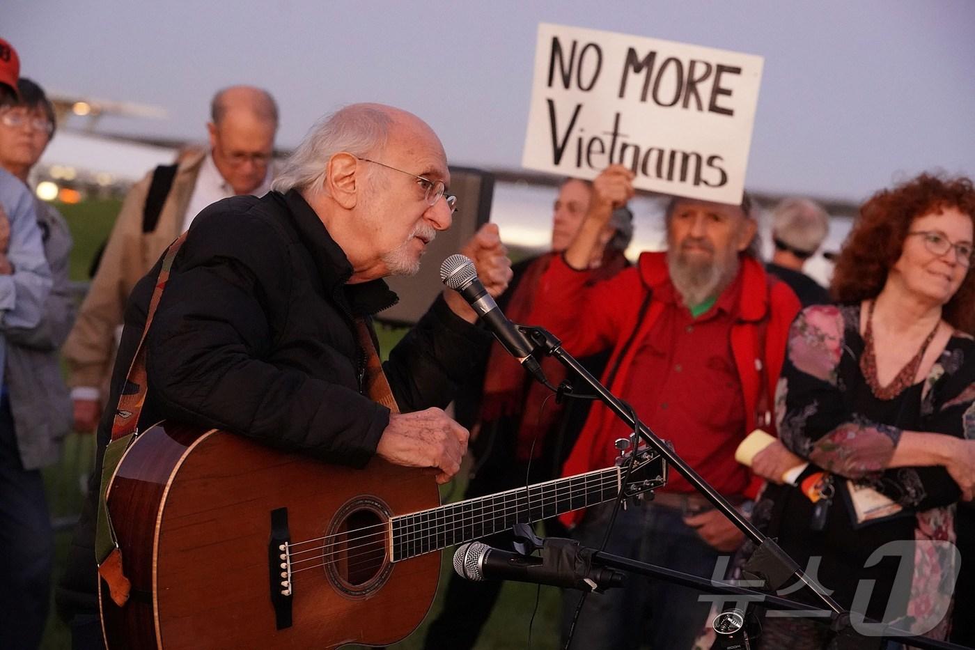 본문 이미지 - 미국 버지니아주(州) 알링턴에서 전설적인 포크송 그룹 '피터 폴 앤 메리'의 창립 멤버, 피터 야로우가 노래하고 있다. 그의 뒤로는 한 남성이 "더 이상의 베트남은 없다＂는 반전 문구가 적힌 팻말을 들고 있다. 2017.10.20/ ⓒ AFP=뉴스1 ⓒ News1 권진영 기자