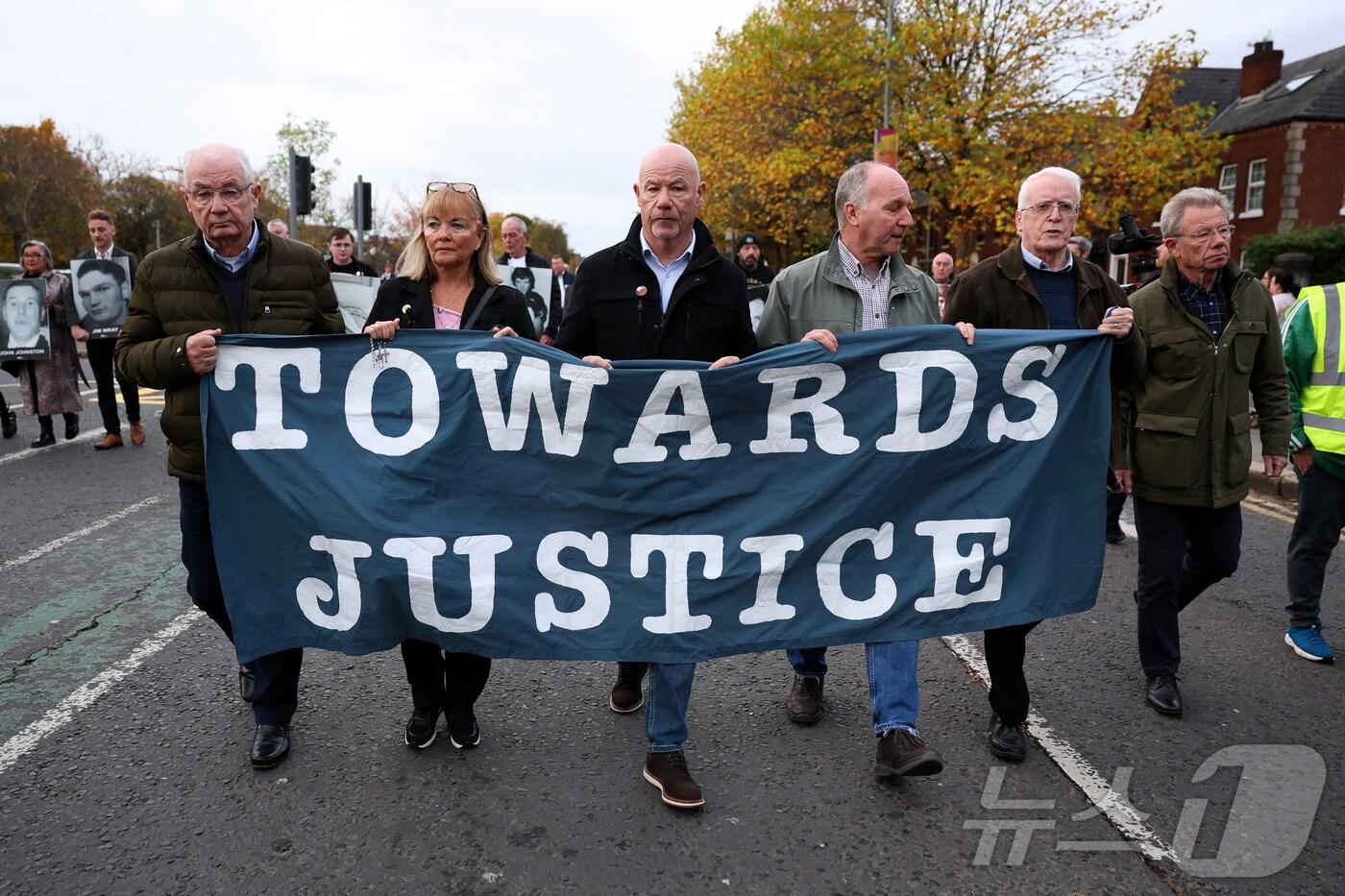 23일(현지시간) 북아일랜드 벨파스트에서 1972년 ‘피의 일요일(Bloody Sunday)’ 희생자 유족들이 모인 가운데, 시민들이 '정의를 향해'라고 쓴 현수막을 들고 있다. 이날 법원은 피의 일요일 사건과 관련해 살인 2건과 살인미수 5건으로 기소된 영국군 참전용사 ‘솔저 F’에 대한 판결을 내렸다. 2025.10.23. ⓒ 로이터=뉴스1