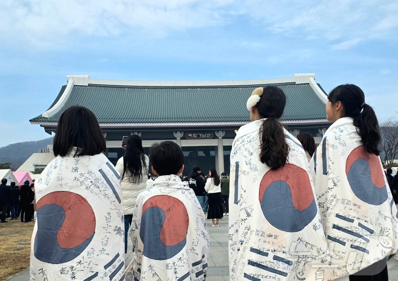 본문 이미지 - 삼일절을 맞아 천안 독립기념관을 방문한 초등학생들이 광복군 태극기를 두르고 겨레의집을 바라보고 있다. 2025.3.1./뉴스1ⓒNews1 이시우 기자