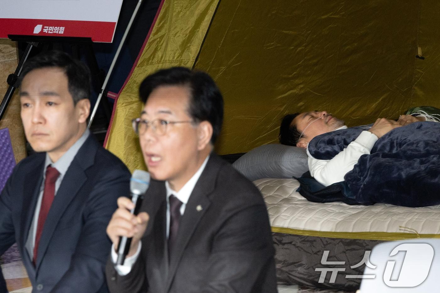 본문 이미지 - 송언석 국민의힘 원내대표가 22일 서울 여의도 국회 로텐더홀에서 열린 최고위원회의에서 모두발언하는 가운데 단식 중인 장동혁 대표가 누워서 휴식을 취하고 있다. 2026.1.22/뉴스1 ⓒ News1 이승배 기자