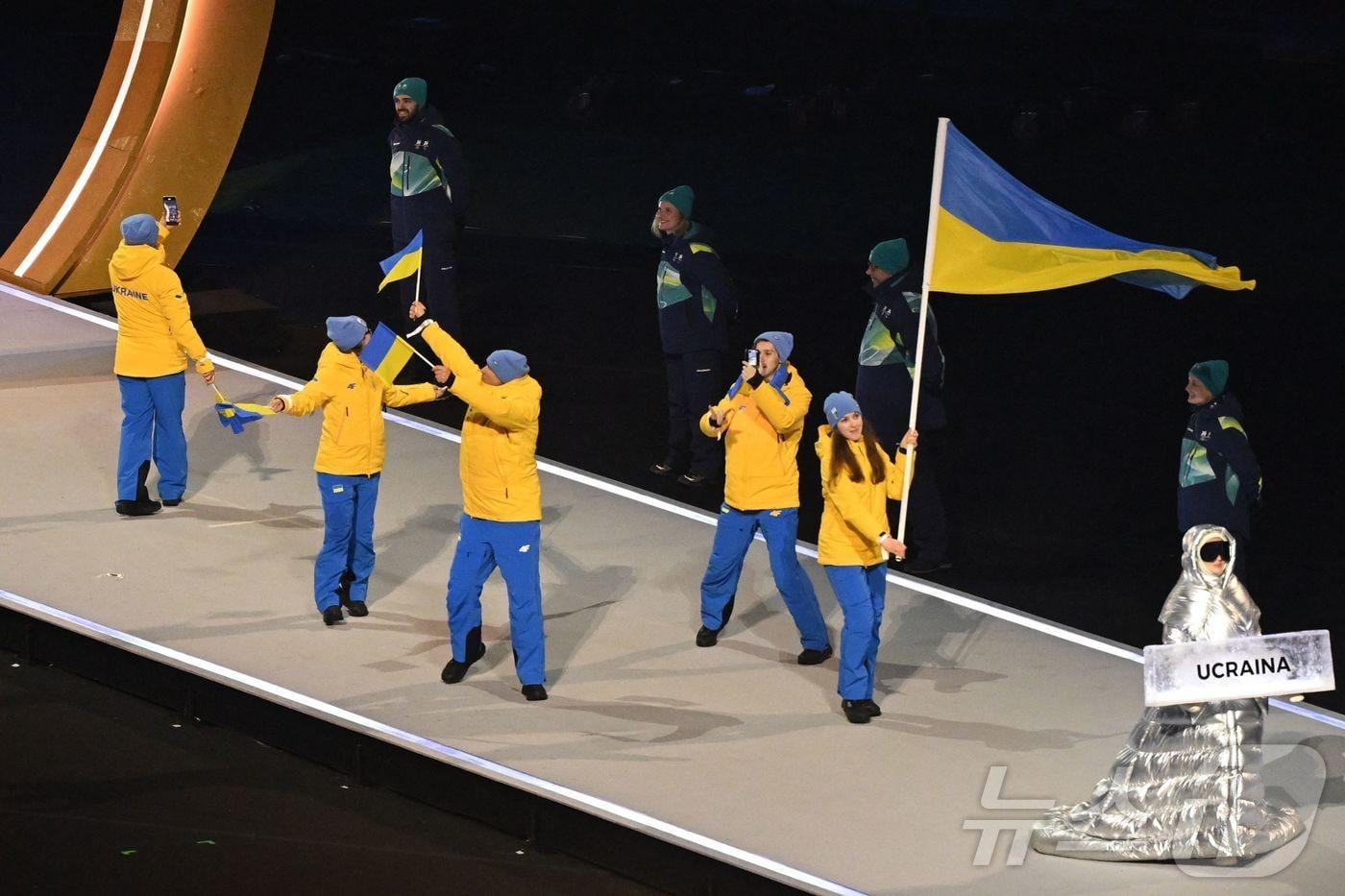 본문 이미지 - 6일(현지시각) 이탈리아 밀라노의 산시로 스타디움에서 열린 2026 밀라노·코르티나담페초 동계올림픽 개막식에서 우크라이나 대표단이 함께 퍼레이드를 하고 있다. 맨 오른쪽 아래  우크라이나 대표단 명판을 든 사람이 아나스타샤 쿠체로바. ⓒ AFP=뉴스1
