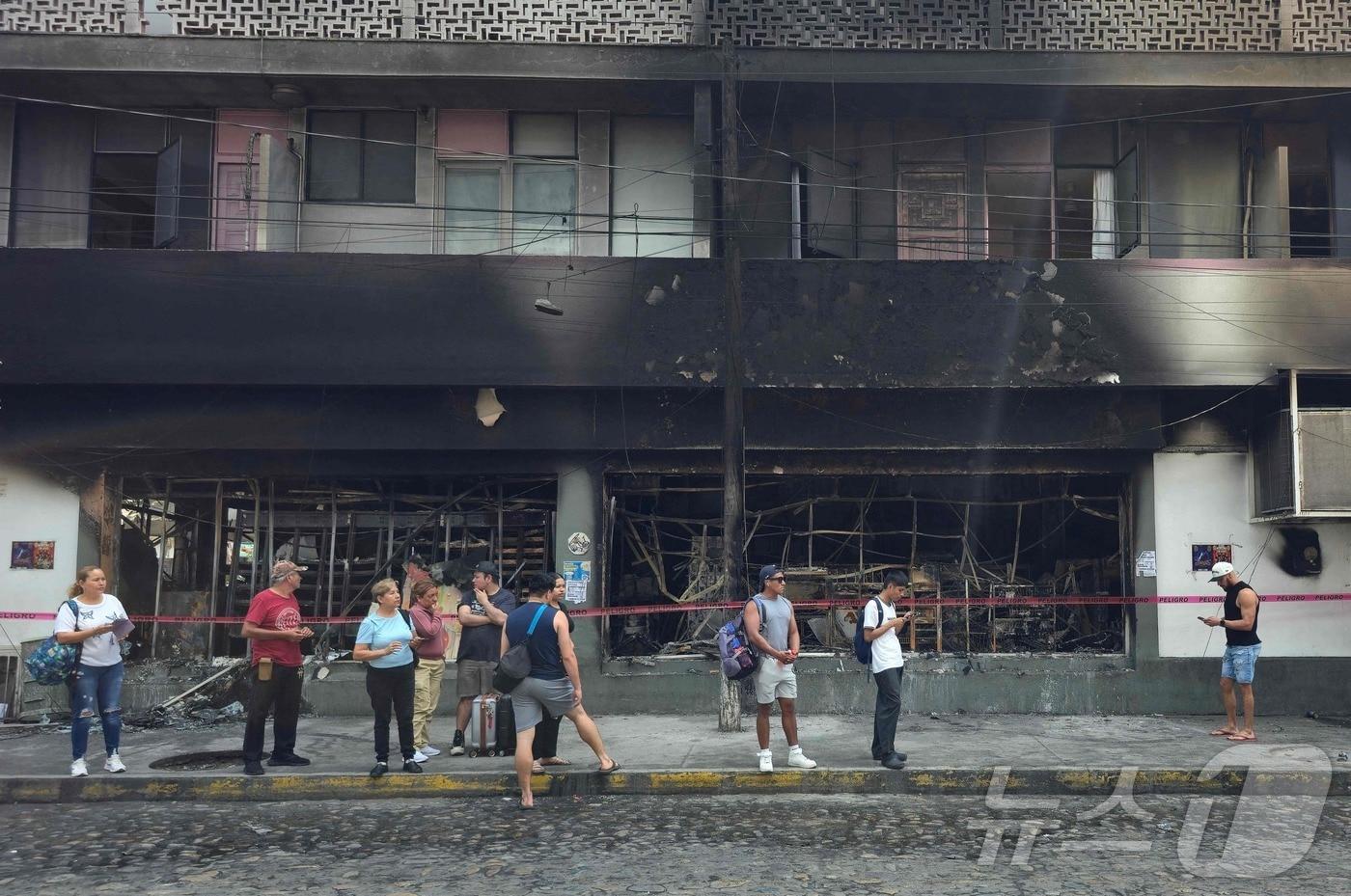 본문 이미지 - 23일(현지시간) 멕시코 할리스코주 푸에르토 바야르타에서 사람들이 불에 탄 건물 옆에 서 있다. 멕시코 당국은 마약왕 사살로 촉발돼 수십 명의 사망자를 낸 유혈 충돌을 진압하기 위해 1만 명의 병력을 배치했다. 2026.02.23 ⓒ AFP=뉴스1