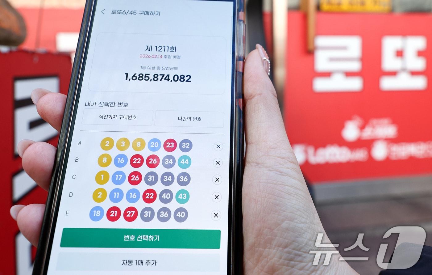 본문 이미지 - 모바일 로또 판매가 시작된 9일 서울 시내의 한 복권 판매점 앞에서 시민이 휴대폰으로 로또를 구매하고 있다. 이용자는 모바일 동행복권 홈페이지에 가입한 뒤 예치금을 충전하면 오전 6시부터 자정까지 복권을 살 수 있다. 시범 운영 기간인 이번 상반기에는 평일에만 회차당 1인 5000원까지 구매할 수 있으며 PC 온라인 구매 금액과 합산해 계산된다. 전체 온라인 판매 규모도 전년도 로또 판매액의 5% 이내로 묶여, 한도에 도달하면 판매가 자동 중단된다. 2026.2.9 ⓒ 뉴스1 최지환 기자