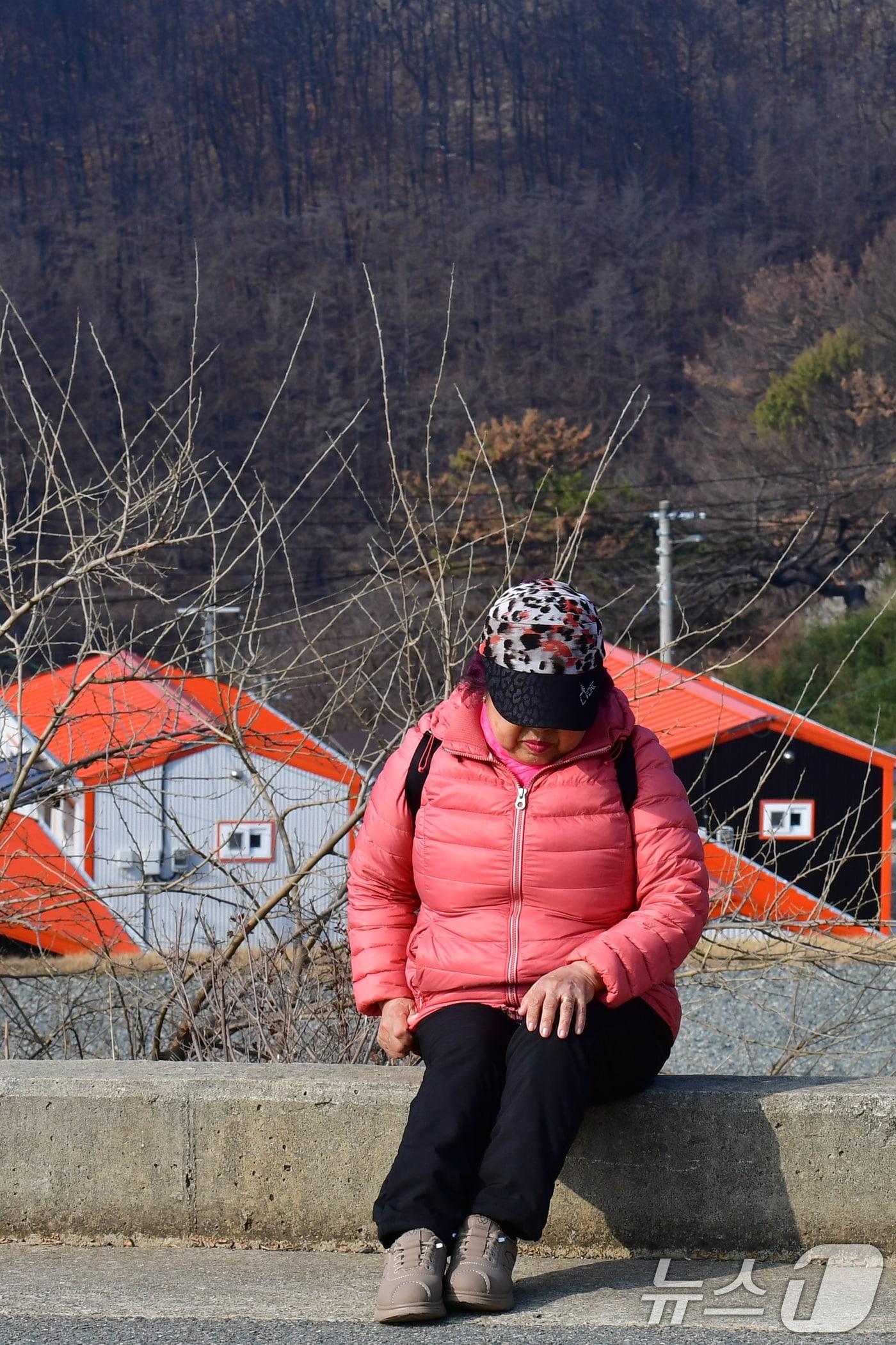 본문 이미지 - 16일 오전 지난해 3월 산불 피해지역인 경북 영덕군 영덕읍 석리 해안 마을(일명 따개비 마을)에서 주민이 버스를 타기 위해 정류장으로 걸어가던 중 도로 옆에 앉아 잠시 쉬고 있다. 주민 뒷편으로 이재민 임시주택과 검개 탄 산이 보인다. 석리는 지난해 3월 22일 의성군에서 발생한 산불이 강풍을 타고 확산되면서 피해가 발생한 지역이다. 산불로 영덕군에서는 약 2만ha(군 전체 면적 약 27%)가 불에 타는 피해가 발생했다. 2026.3.16 ⓒ 뉴스1 최창호 기자