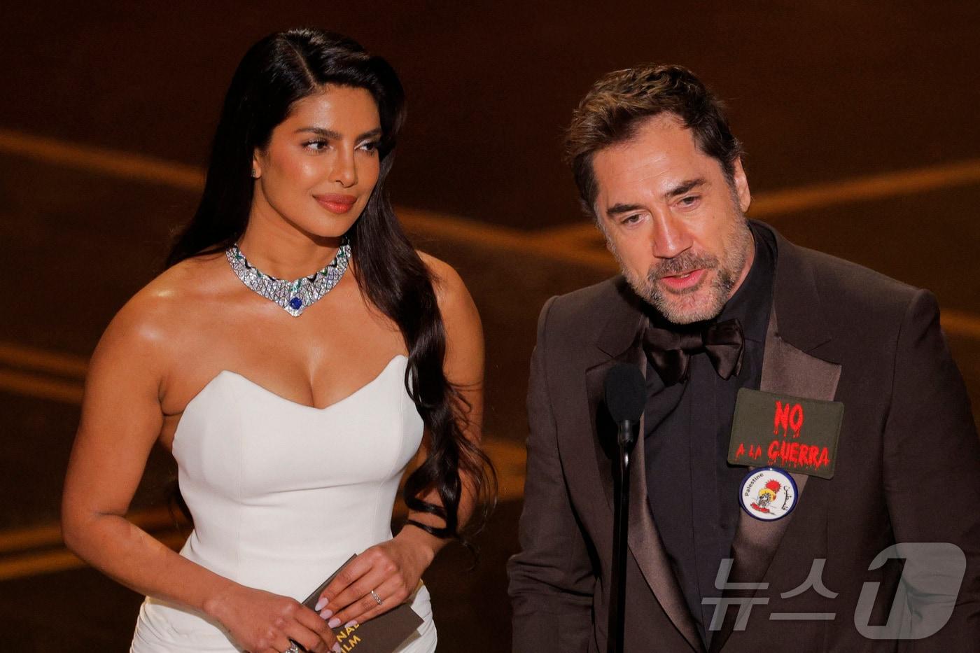 본문 이미지 - 15일(현지시간) 미국 캘리포니아주 로스앤젤레스(LA) 할리우드에서 열린 제98회 아카데미 시상식에서 배우 하비에르 바르뎀(오른쪽)이 '전쟁을 멈춰라'(NO A LA GUERRA)라는 스페인어 문구를 가슴에 착용하고 배우 프리양카 초프라와 함께 무대에 올라 발언하고 있다. 2026.03.15. ⓒ 로이터=뉴스1