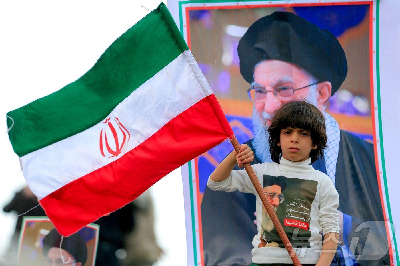 본문 이미지 - 이란 국기를 든 소년. 뒤에는 미국의 공습을 받고 사망한 이란 전 최고 지도자 아야톨라 알리 하메네이.  ⓒ AFP=뉴스1