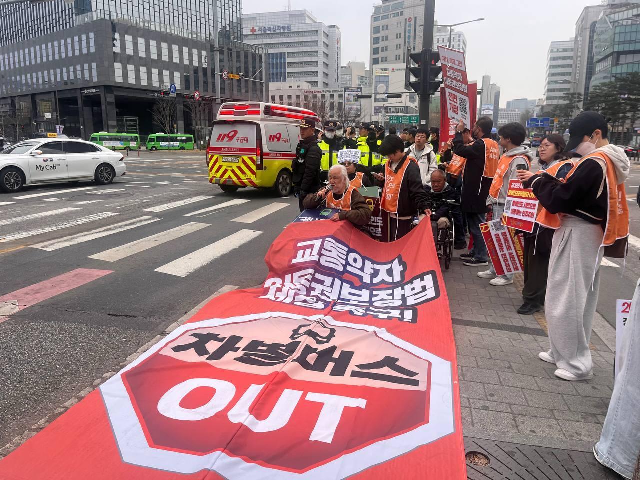 본문 이미지 - 30일 오전 전국장애인차별철폐연대가 서울 서대문구 서대문사거리 버스 정류장에서 대형 현수막을 펼치고 시위하는 모습(전장연 제공)