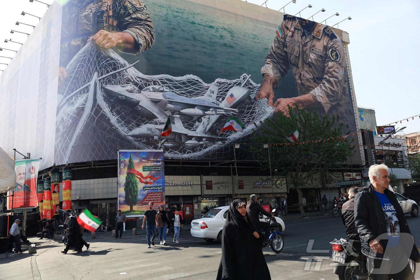 본문 이미지 - 16일(현지시간) 이란 테헤란의 한 건물 앞을 시민들이 지나가고 있다. 건물 외벽에는 반미 선전물이 게시돼 있다. 2026.04.16 ⓒ 로이터=뉴스1