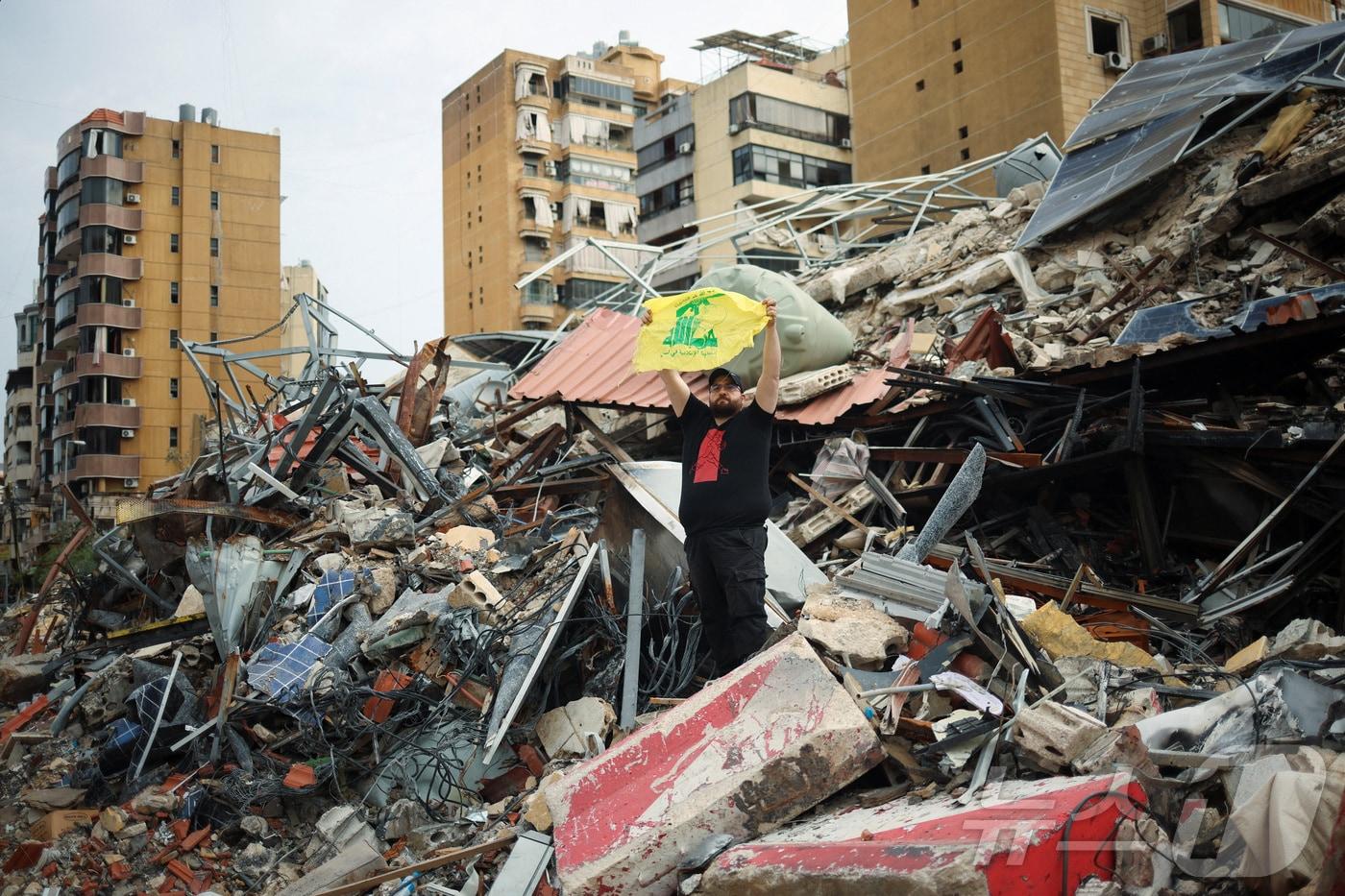 본문 이미지 - 17일(현지시간) 레바논 베이루트의 한 건물 잔해에서 주민이 현지 무장정파 헤즈볼라의 깃발을 들고 있다. 2026.04.17 ⓒ 로이터=뉴스1