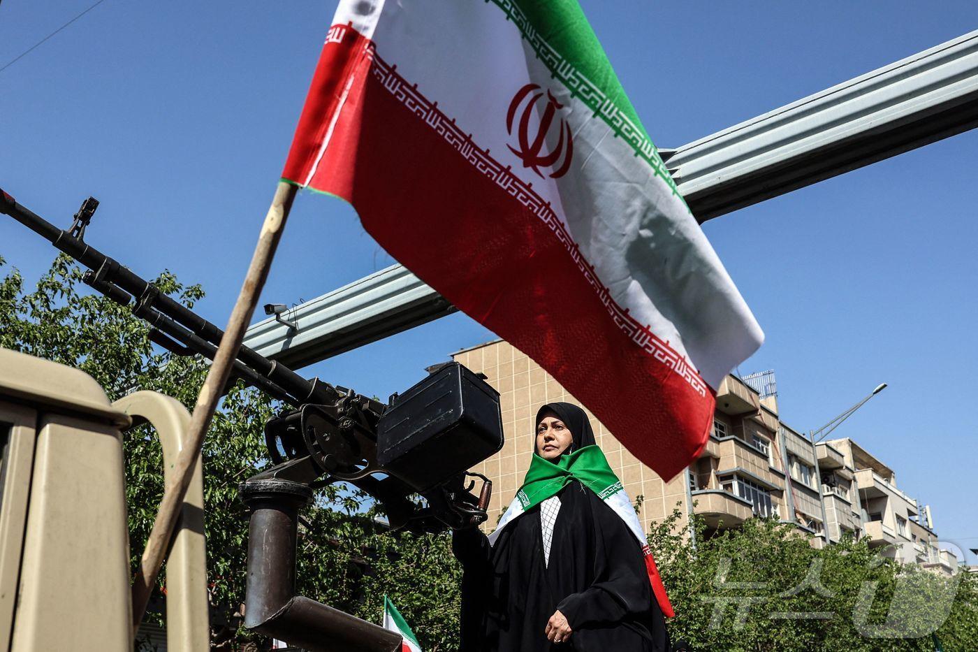 본문 이미지 - 17일(현지시간) 이란 수도 테헤란에서 열린 '희생된 소녀들' 집회에 참가한 한 이란 여성이 차량 뒤에 장착된 중기관총 앞에 서 있다. 이 집회는 중동 전쟁 중 희생된 여성들을 추모하기 위해 열렸다. 2026.04.18. ⓒ AFP=뉴스1