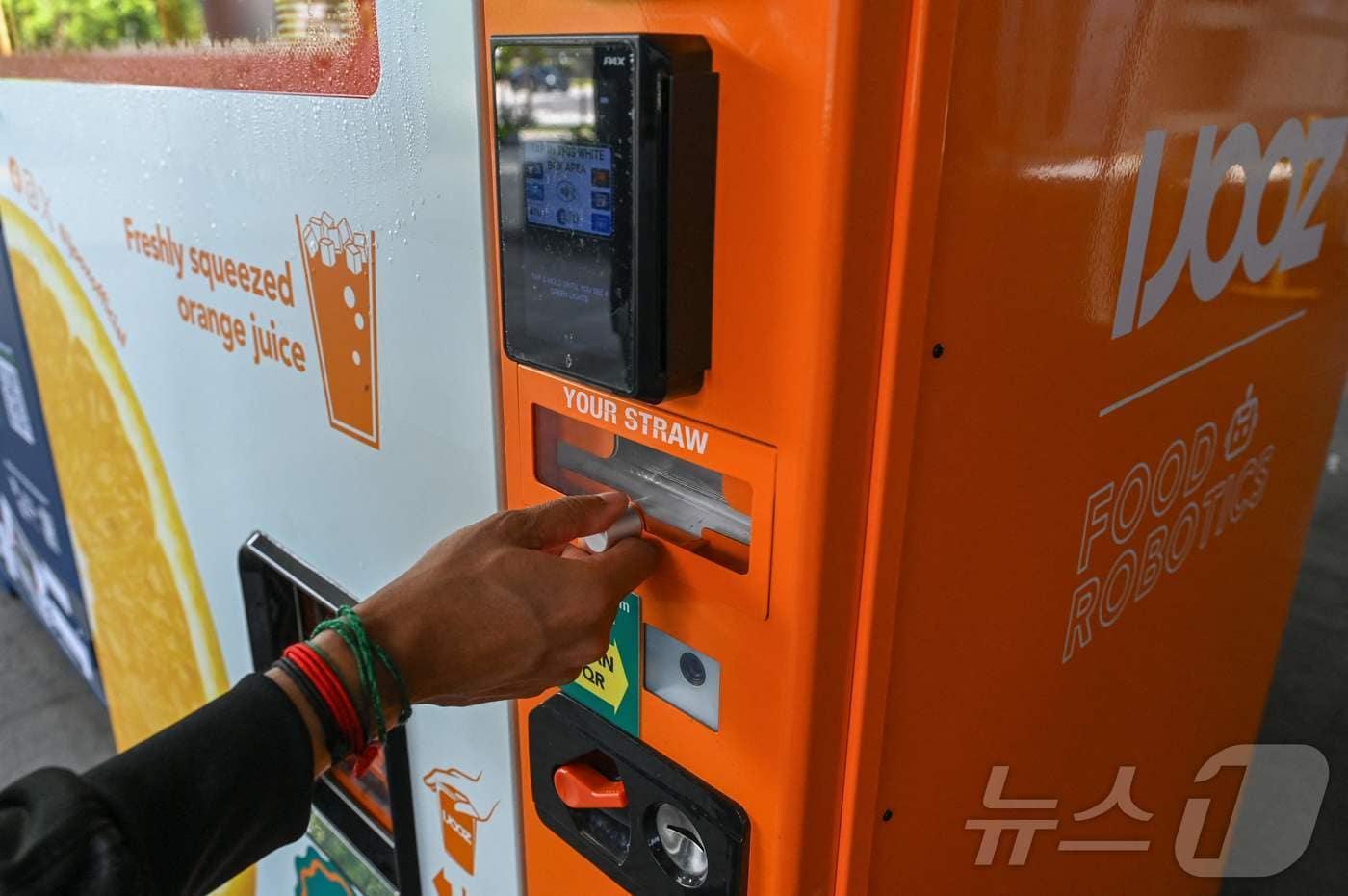 본문 이미지 - 28일(현지시간) 싱가포르의 한 쇼핑몰 앞에서 한 남성이 아이주스(IJOOZ)사의 오렌지 주스 자판기 빨대 칸에 손을 뻗고 있다. CNN에 따르면 전날(27일) 프랑스 국적의 18세 청소년이 오렌지 주스 자판기의 빨대를 핥은 뒤 다시 넣은 혐의로 기물 손괴 및 공공 불법 방해 혐의로 기소됐다. 2026.04.28. ⓒ AFP=뉴스1