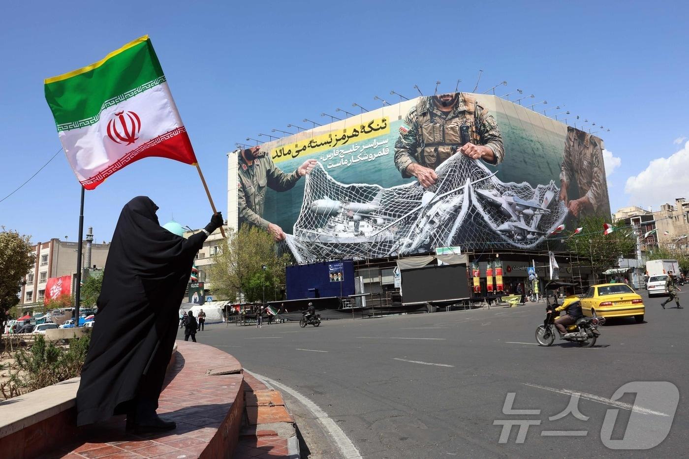 본문 이미지 - 5일(현지시간) 이란 수도 테헤란 엔켈랍 광장에서 한 여성이 "호르무즈 해협은 여전히 ​​폐쇄되어 있다＂라는 문구가 적힌 광고판 옆에 서서 이란 국기를 들고 있다. 2026.04.05. ⓒ AFP=뉴스1