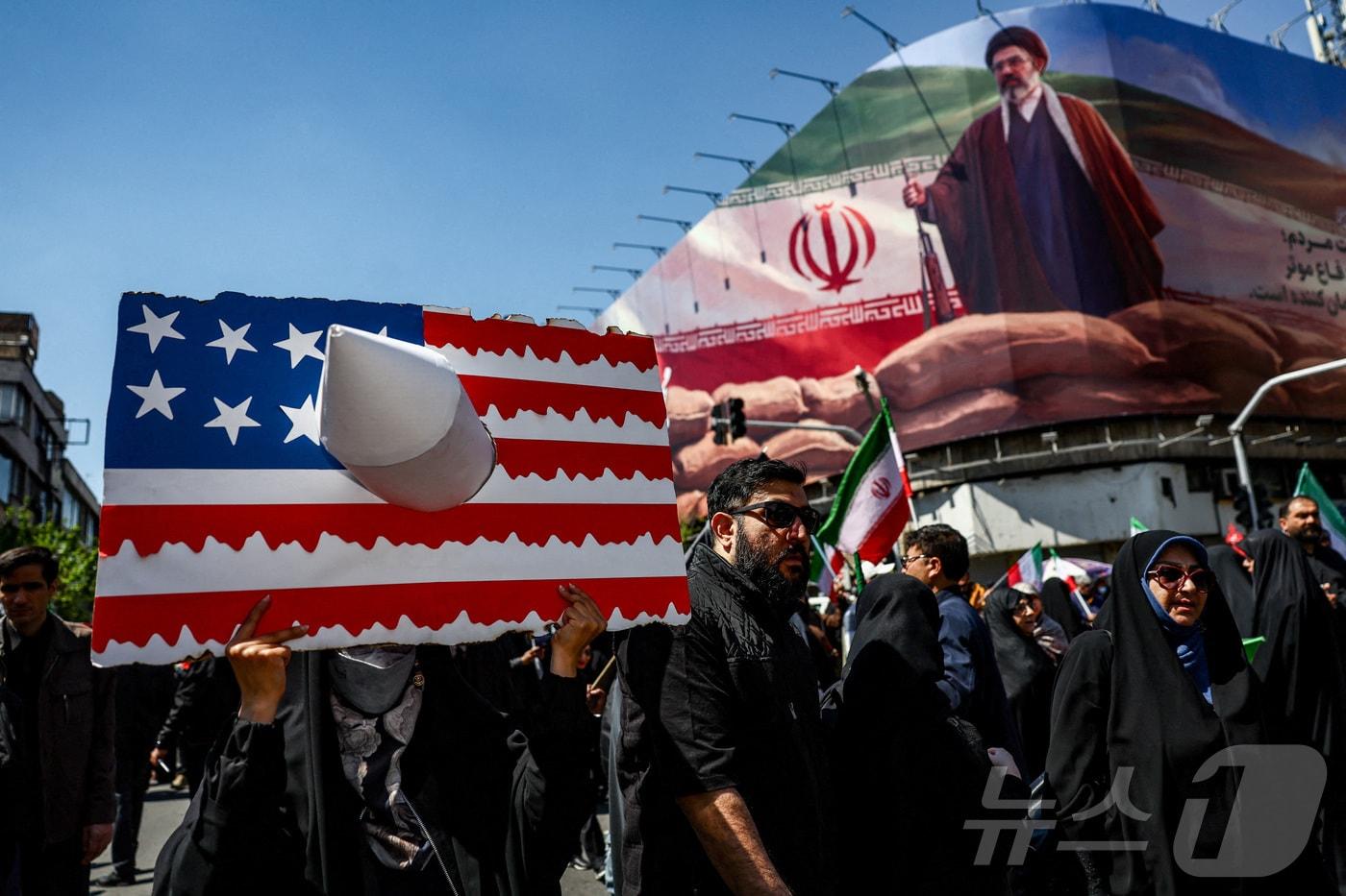 본문 이미지 - 이란 테헤란에서 열린 아야톨라 알리 하메네이 사망 40일 추모식. 미사일로 구멍난 성조기를 든 참가자들 뒤로 새 최고지도자 모즈타바 하메네이의 사진이 보인다. 2026.04.09 ⓒ 로이터=뉴스1