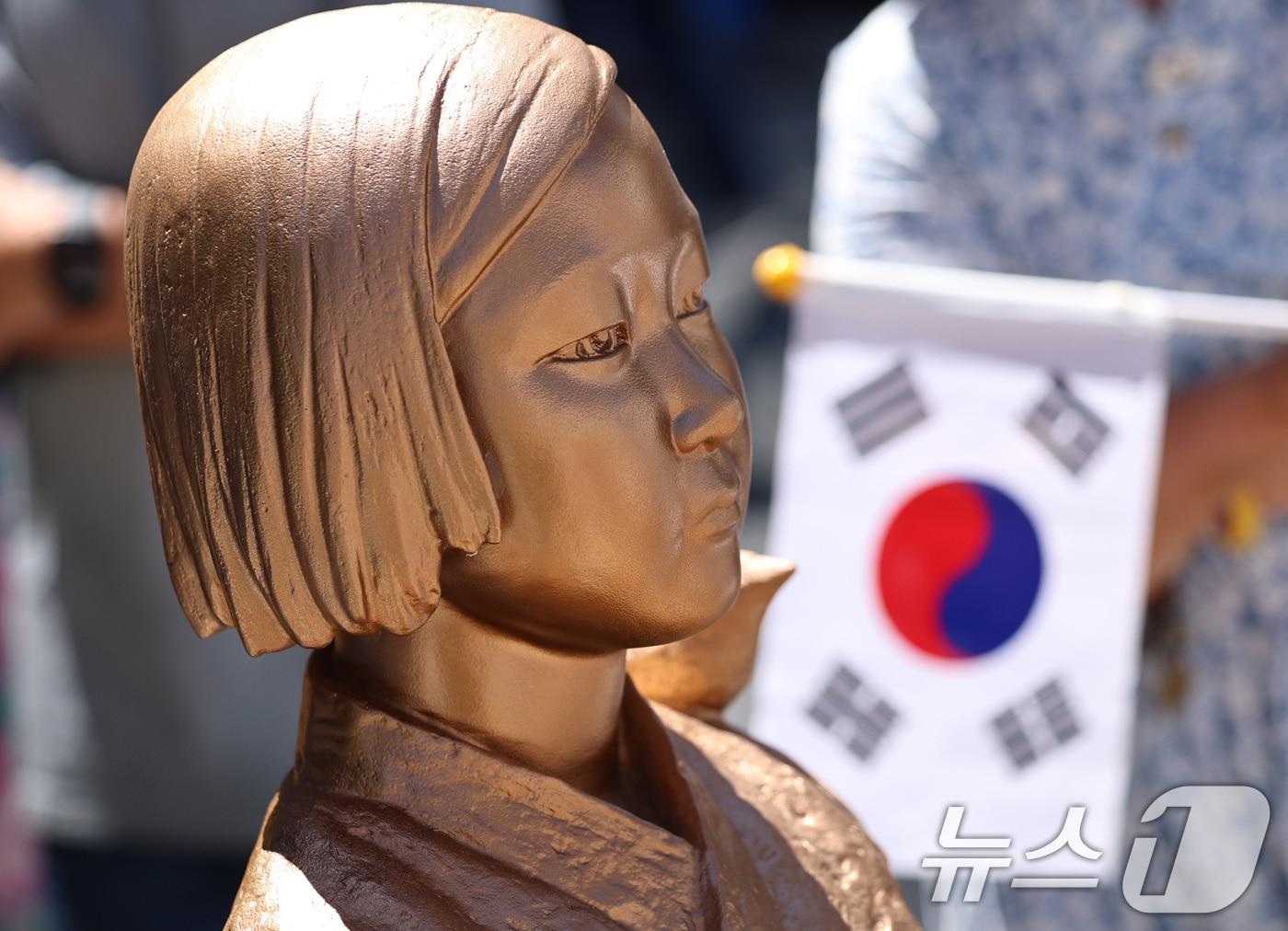본문 이미지 - 서울 종로구 옛 주한일본대사관 앞에서 열린 제14차 세계일본군 '위안부' 기림의 날 맞이 세계연대집회에서 태극기를 든 한 참가자가 평화의 소녀상 곁에 서 있다. 2026.8.12 ⓒ 뉴스1 구윤성 기자