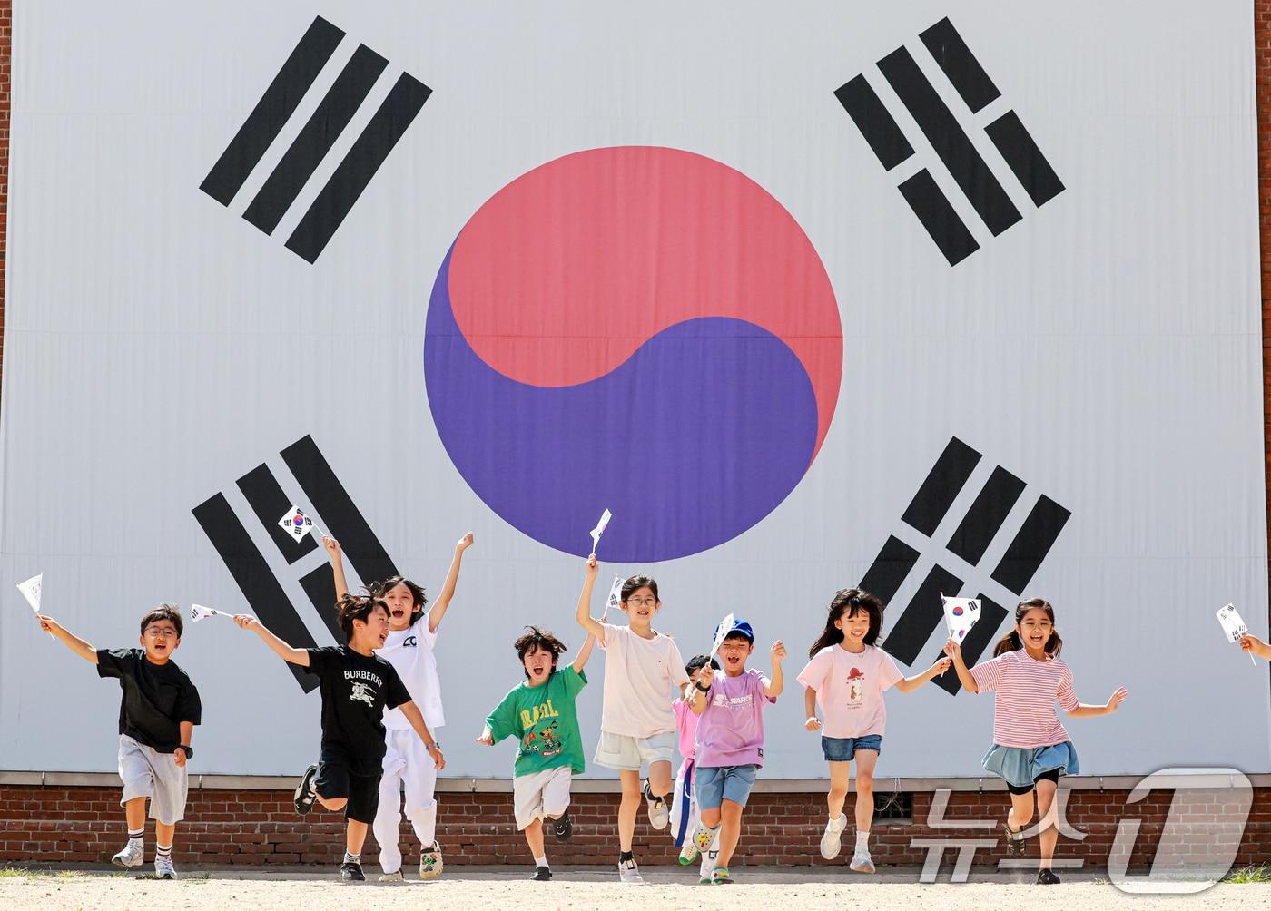 본문 이미지 - 제81주년 광복절을 하루 앞둔 14일 서울 서대문구 서대문형무소역사관 대형 태극기 앞에서 어린이들이 직접 만든 태극기를 흔들며 힘차게 뛰고 있다. 2026.8.14 ⓒ 뉴스1 이호윤 기자