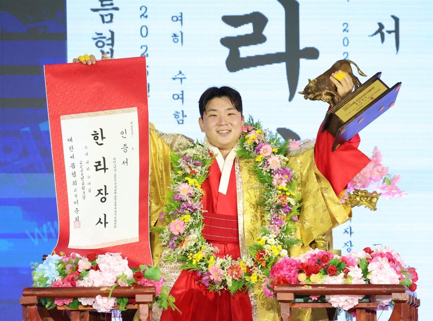 본문 이미지 - ‘위더스제약 2026 민속씨름 문경오미자장사씨름대회’ 한라장사에 등극한 김무호(울주군청)가 장사꽃가마를 타고 장사인증서와 황소트로피를 들고 기념사진을 찍고 있다.(대한씨름협회 제공)