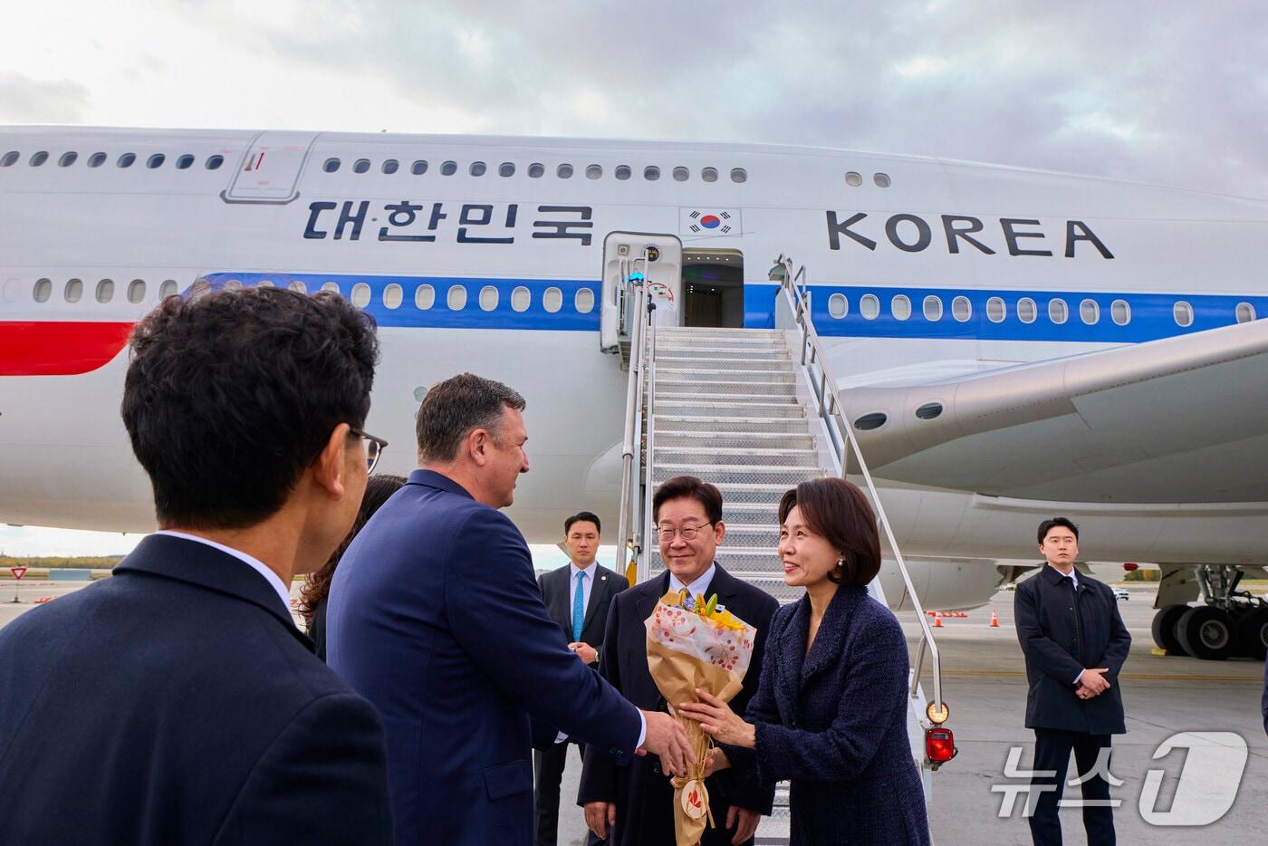본문 이미지 - 이재명 대통령과 김혜경 여사가 26일(현지시간) 순방 귀국길 경유지인 미국 앵커리지에 도착해 환영객에게 꽃다발을 전달 받고 있다. (공동취재) 2026.9.27 ⓒ 뉴스1 허경 기자